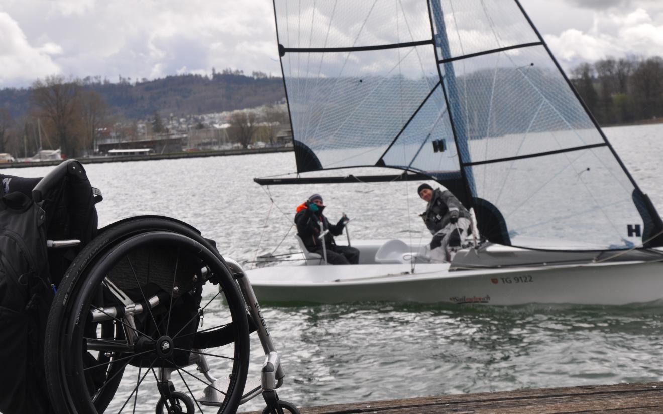Leerer Rollstuhl am Steg mit Blick auf angepasstes Segelboot auf dem Wasser